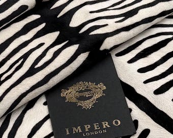 Op de afbeelding: Een stuk stof met een zwart-wit zebrastreeppatroon. Bovenop de stof ligt een zwart vierkant label met de gouden tekst "IMPERO LONDON" en een goudkleurig sierlijk embleem boven de merknaam, gecentreerd op het label.