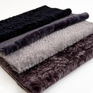 Op de afbeelding: Vier stofstalen in grijstinten en bruin zijn op elkaar gestapeld. De kleuren variëren van zwart tot donkergrijs, lichtgrijs en een diep bruin. De stof heeft een zachte, pluche uitstraling. Op een witte achtergrond.