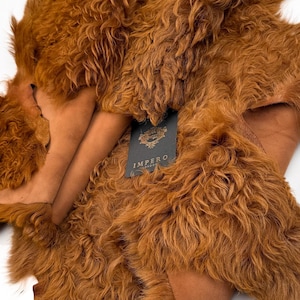 May include: A close-up shot of a brown sheepskin rug with a curly, textured surface. The rug has a rich, warm color and a soft, inviting appearance. A tag with the word "IMPERO" is visible.