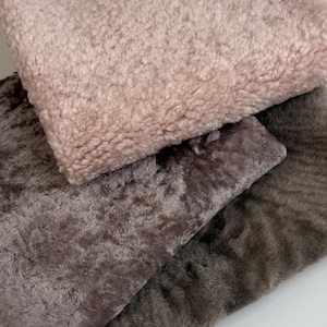 Op de afbeelding: Drie zachte, getextureerde stoffen in tinten roze, bruin en grijs zijn op elkaar gestapeld. Het bovenste stuk is lichtroze, het middelste is donkerder bruin en het onderste is grijsbruin. De stoffen zien er zacht en luxueus uit.