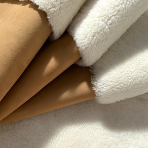 May include: Close-up of a white faux fur fabric with a brown suede fabric underneath. The faux fur is soft and fluffy, while the suede is smooth and textured.