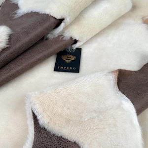 May include: Close-up of a white sheepskin rug with a brown leather backing. The rug is folded in half, showing both the fur and leather sides. The rug has a label that reads "Impero London".