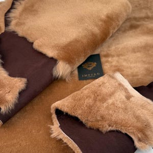 Könnte beinhalten: Nahaufnahme von mehreren Stücken hellbraunem Kunstfellstoff mit dunkelbraunem Rücken. Das Fell hat eine weiche, flauschige Textur. Die Kanten des Stoffes sind sichtbar und zeigen den Kontrast zwischen Fell und Rückseite. Ein Etikett mit dem Wort "IMPERO" ist sichtbar.