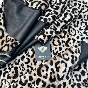 May include: A close-up of a beige and black leopard print fabric with a black leather backing. The fabric is folded over itself, and the label "Impero London" is visible.