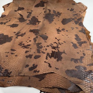 May include: A stack of brown and black snakeskin leather hides. The leather has a textured pattern and a mottled appearance.