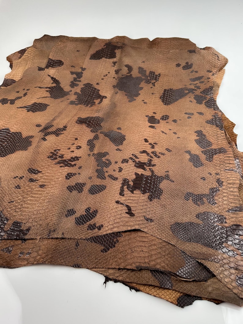 May include: A stack of brown and black snakeskin leather hides. The leather has a textured pattern and a mottled appearance.