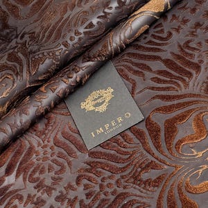 Könnte beinhalten: Nahaufnahme von braunem Lederstoff mit geprägtem floralem Muster. Das Leder ist gefaltet und zeigt die Textur und Tiefe des Designs. Ein Etikett mit dem Text "IMPERO LONDON" ist sichtbar.
