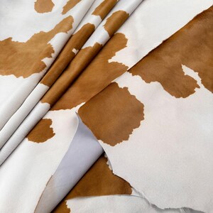 May include: A close-up of a cowhide leather with a white background and brown patches. The leather is folded and appears to be soft and pliable. The brown patches vary in size and shape, creating a natural pattern.
