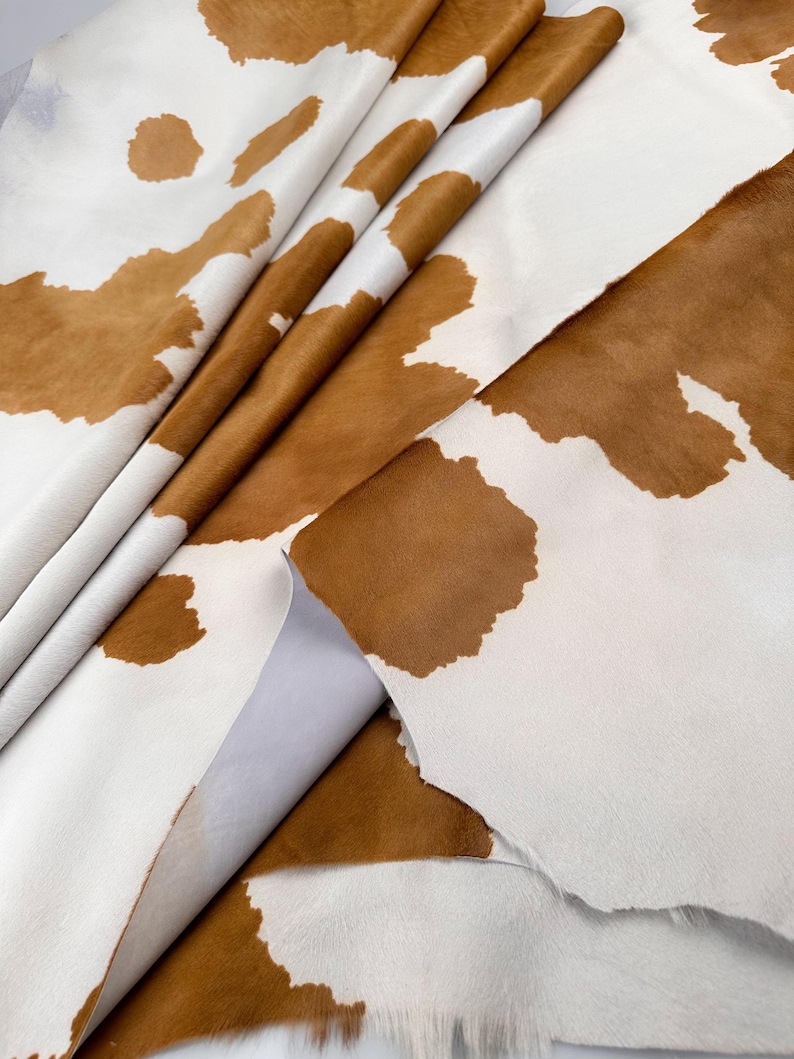 May include: A close-up of a cowhide leather with a white background and brown patches. The leather is folded and appears to be soft and pliable. The brown patches vary in size and shape, creating a natural pattern.