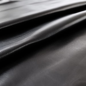 May include: Close-up of a smooth, black leather fabric with a subtle sheen. The fabric is folded and draped, showcasing its texture and drape.