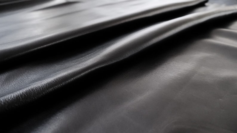 May include: Close-up of a smooth, black leather fabric with a subtle sheen. The fabric is folded and draped, showcasing its texture and drape.