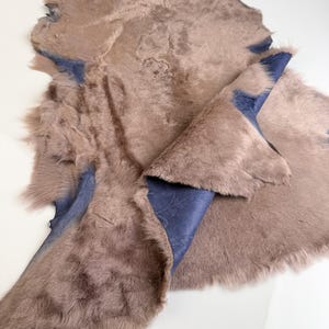 K&ouml;nnte beinhalten: Ein gegerbtes, pelziges Fell mit einer blau gemusterten Unterseite. Das Fell hat eine ged&auml;mpfte braune Farbe mit einer weichen Textur. Die blaue Unterseite ist dort sichtbar, wo das Fell umgeschlagen ist und ein detailliertes Muster zeigt. Das Gesamtbild deutet auf einen nat&uuml;rlichen, handgefertigten Artikel hin.