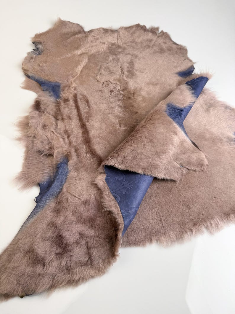 K&ouml;nnte beinhalten: Ein gegerbtes, pelziges Fell mit einer blau gemusterten Unterseite. Das Fell hat eine ged&auml;mpfte braune Farbe mit einer weichen Textur. Die blaue Unterseite ist dort sichtbar, wo das Fell umgeschlagen ist und ein detailliertes Muster zeigt. Das Gesamtbild deutet auf einen nat&uuml;rlichen, handgefertigten Artikel hin.