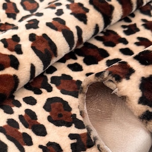 May include: Close-up of a plush fabric with a leopard print pattern. The fabric features a beige background with brown and black spots. The material is folded, revealing a smooth, light brown backing.