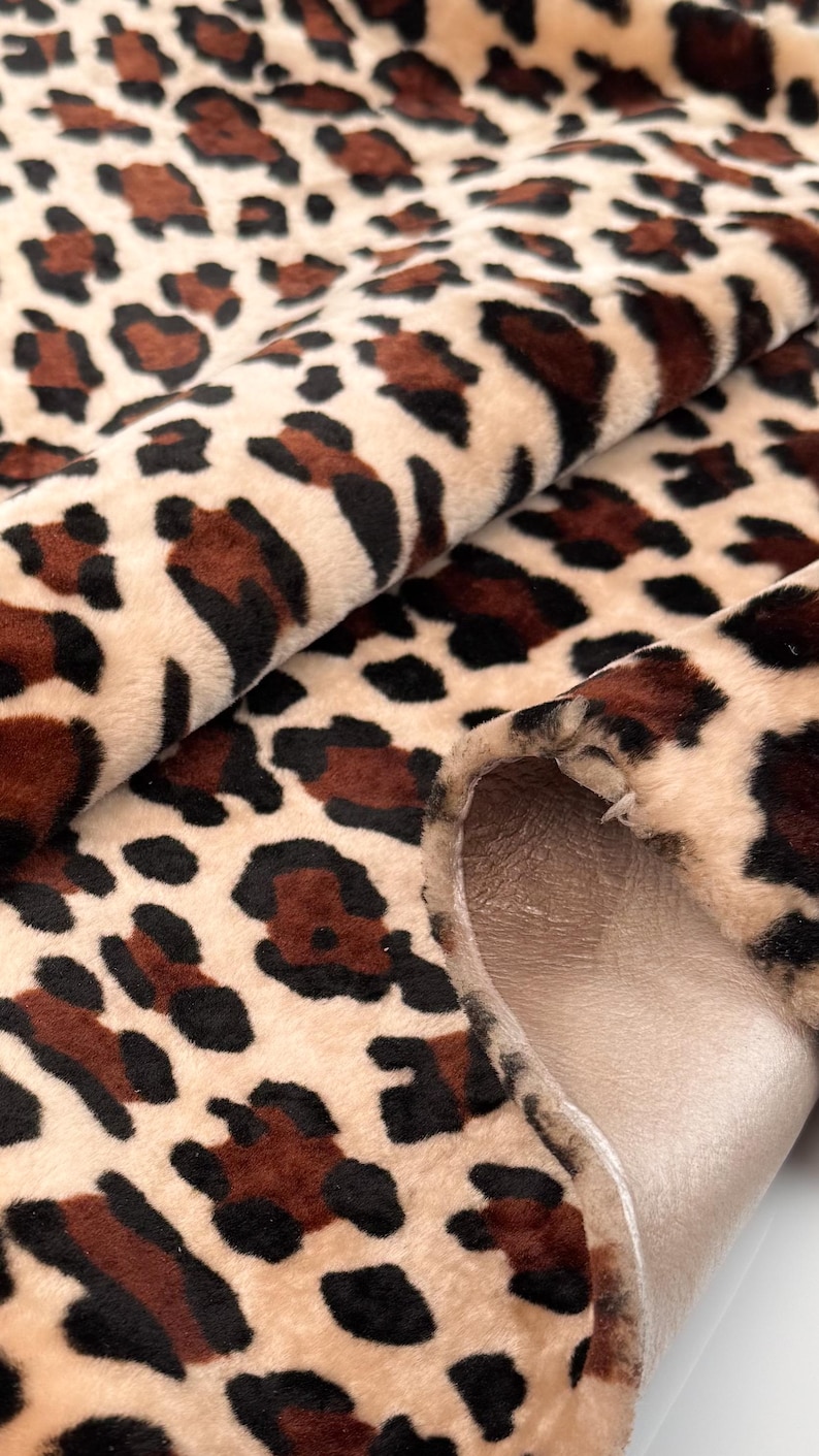 May include: Close-up of a plush fabric with a leopard print pattern. The fabric features a beige background with brown and black spots. The material is folded, revealing a smooth, light brown backing.
