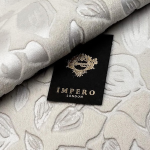 May include: Close-up of a cream-colored fabric with a raised floral pattern. The fabric is folded, revealing its texture and design. A black tag with gold lettering that reads "IMPERO LONDON" is placed on the fabric.