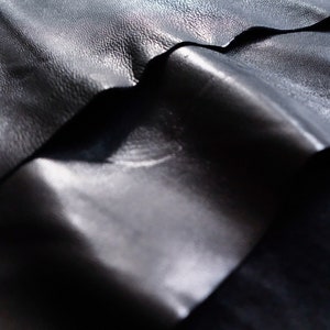 May include: Close-up of three pieces of black leather stacked on top of each other. The leather has a smooth, shiny surface and a slightly textured grain.