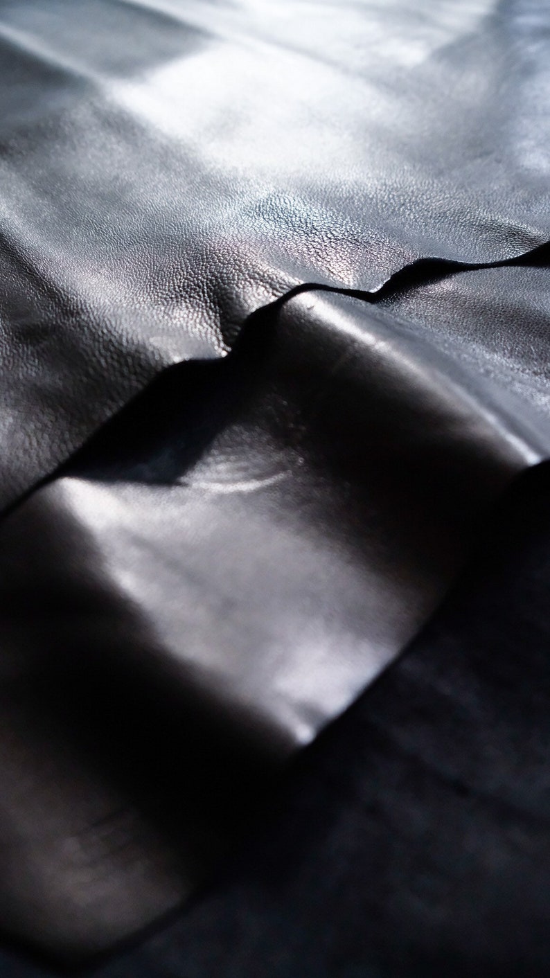 May include: Close-up of three pieces of black leather stacked on top of each other. The leather has a smooth, shiny surface and a slightly textured grain.