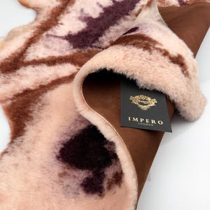 Könnte beinhalten: Ein Schaffellteppich mit einem einzigartigen Muster in Rosa-, Braun- und Burgundertönen. Der Teppich hat eine weiche, flauschige Textur und eine braune Lederunterseite. Ein schwarzes Etikett mit goldener Schrift lautet "IMPERO LONDON".