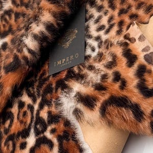 以下が含まれることがあります： 茶色、黒、クリーム色の斑点模様のヒョウ柄生地のクローズアップ。生地は柔らかく、質感があります。「IMPERO LONDON」の文字が入った黒いタグが生地に付いています。
