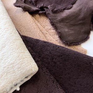 May include: Close-up of various sheepskin hides in shades of cream, tan, and deep brown. The textures vary from smooth leather to thick, fluffy fleece. The hides are layered, showcasing the different textures and colors of the natural material.