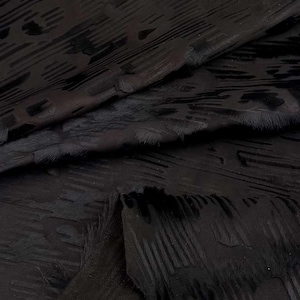 May include: A piece of black leather featuring a textured surface with a raised, repeating pattern. The material displays a combination of smooth, matte sections and areas with fine, hair-like fibres that create a distinct visual depth and contrast.