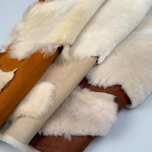 K&ouml;nnte beinhalten: Vier Schaffellteppiche in verschiedenen Farben, darunter Wei&szlig;, Beige und Braun. Die Teppiche sind gefaltet und &uuml;bereinander gestapelt.