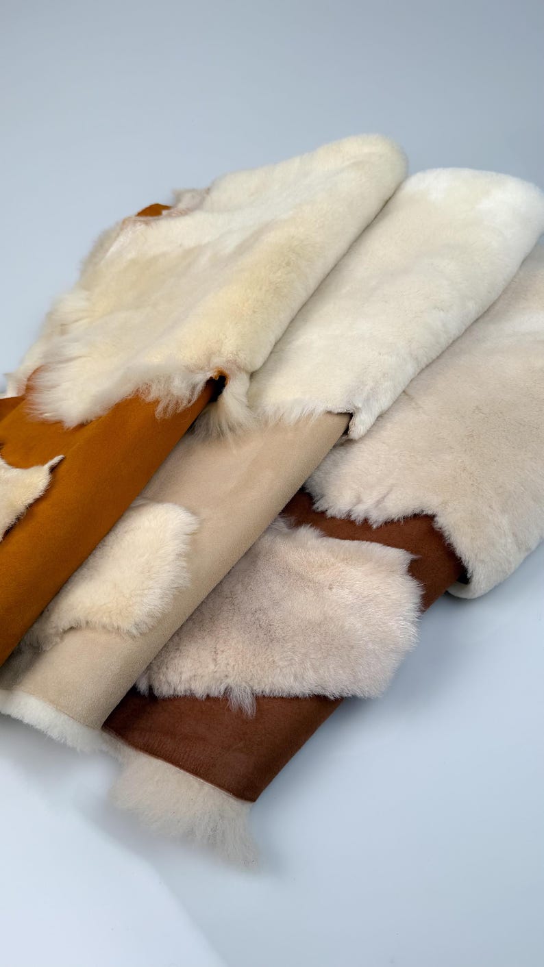 K&ouml;nnte beinhalten: Vier Schaffellteppiche in verschiedenen Farben, darunter Wei&szlig;, Beige und Braun. Die Teppiche sind gefaltet und &uuml;bereinander gestapelt.