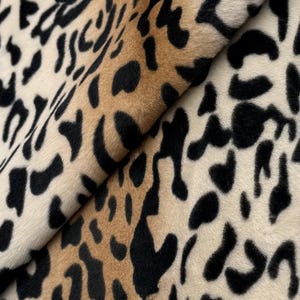 May include: A soft, plush fabric featuring a classic leopard print pattern. The material displays a repeating design of black spots against a background that transitions from a warm, golden tan to a lighter, creamy off-white colour throughout.