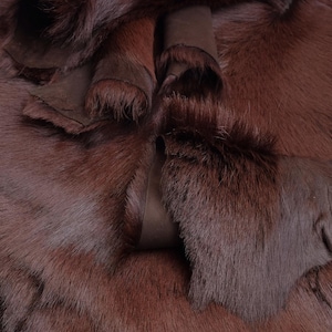 Puede incluir: Una colección de pieles de animal de color marrón oscuro con pelaje denso y suave en un lado y una superficie de cuero marrón mate y lisa en el reverso. Las pieles están superpuestas y dobladas, mostrando la textura del pelo y el reverso.