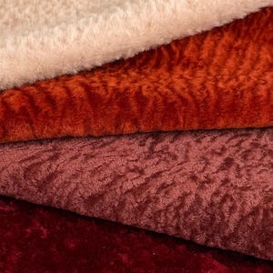 May include: Close-up of four different textured fabrics in shades of cream, orange, burgundy, and deep red. The fabrics have a soft, plush appearance, with visible fibers and a slightly uneven surface. The image highlights the textures and colors of the materials.