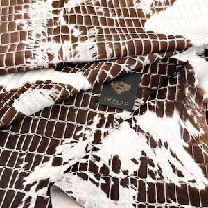 Könnte beinhalten: Nahaufnahme von braunem und weißem Rindsleder mit reptilienartigem Muster. Das Fell ist gefaltet und zeigt die Textur und Farbvariationen. Ein schwarzes Etikett mit goldenem Text lautet "IMPERO LONDON".