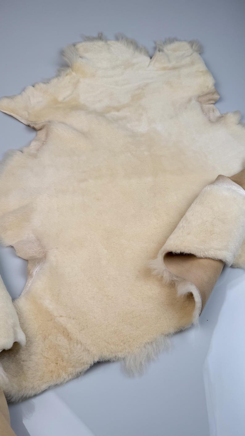 K&ouml;nnte beinhalten: Ein cremefarbener Schaffellteppich mit weicher, flauschiger Textur. Der Teppich ist in der Mitte gefaltet, wodurch die braune Lederunterseite sichtbar wird.