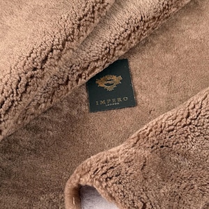 May include: Close-up of a brown sheepskin rug with a label that reads "Impero London". The rug has a soft, fluffy texture.