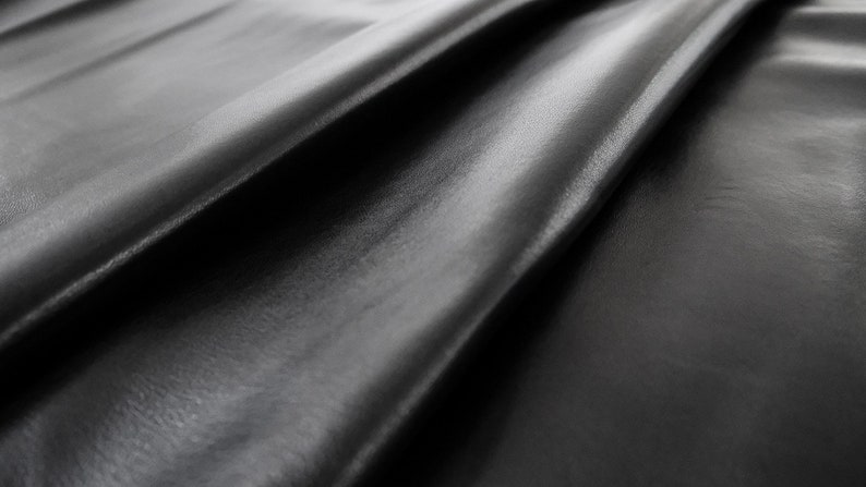 May include: Close-up of a smooth, black fabric with a soft, silky texture. The fabric is draped in folds, creating a luxurious and elegant look.