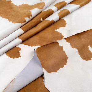May include: Close-up of a cowhide leather material. The leather has a white background with irregular brown patches. The material is folded over itself, revealing the underside of the leather.