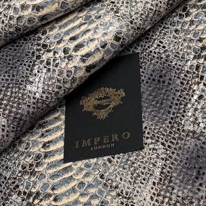 Könnte beinhalten: Nahaufnahme eines strukturierten Schlangenmuster in Grau-, Silber- und Beigetönen. Ein schwarzes Etikett mit goldfarbenem Schriftzug lautet "IMPERO LONDON". Das Schlangenmuster hat ein schuppenartiges Design.