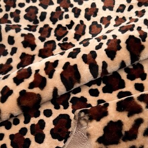 May include: Close-up of a plush fabric with a leopard print pattern. The fabric features a beige background with dark brown and black spots. The material is folded, revealing a smooth, light beige underside.