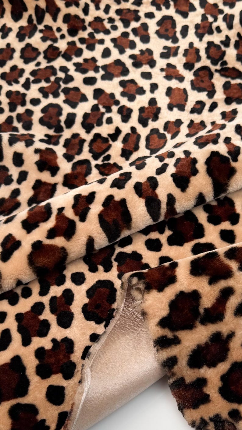 May include: Close-up of a plush fabric with a leopard print pattern. The fabric features a beige background with dark brown and black spots. The material is folded, revealing a smooth, light beige underside.