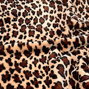 May include: Close-up of a plush fabric with a leopard print pattern. The fabric has a beige background with dark brown and black spots. The material is soft and textured, with visible folds and shadows.