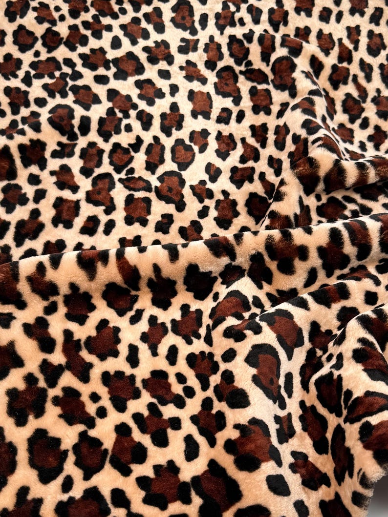 May include: Close-up of a plush fabric with a leopard print pattern. The fabric has a beige background with dark brown and black spots. The material is soft and textured, with visible folds and shadows.