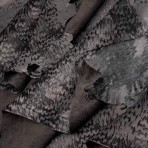 Puede incluir: Primer plano de varios trozos de cuero gris oscuro con una superficie texturizada y estampada. El cuero tiene un aspecto moteado con áreas de tonos más claros y oscuros, y algunas piezas están dobladas.