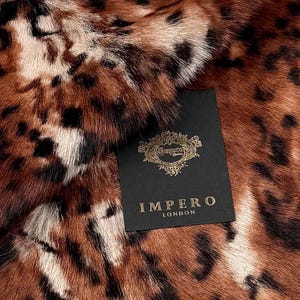 Puede incluir: Una piel animal texturizada con un patrón de manchas marrones, negras y blancas. Se adjunta una etiqueta rectangular negra que presenta un emblema dorado ornamentado en la parte superior y el texto IMPERO LONDON en letras doradas.