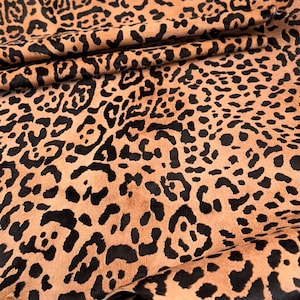 May include: Close-up of a tan and black leopard print hide. The hide has a soft, textured appearance with irregular black spots scattered across a tan background. The material is folded, revealing the underside.