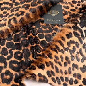 Puede incluir: Primer plano de una piel de leopardo. La piel presenta un patrón de manchas negras sobre un fondo marrón y beige. Una etiqueta negra con texto dorado que dice "IMPERO LONDON" es visible.
