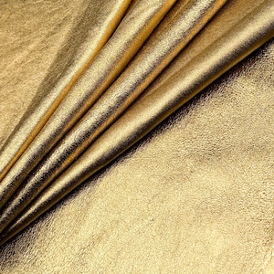 May include: Close-up of a folded sheet of gold-colored fabric. The fabric has a textured, slightly distressed appearance, with visible folds and creases. The lighting highlights the metallic sheen and variations in the surface.