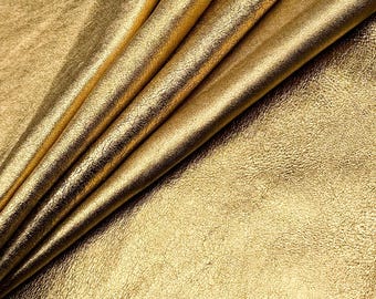 Może przedstawiać: Zbliżenie na złożony arkusz złotej tkaniny. Tkanina ma teksturowany, lekko postarzany wygląd, z widocznymi fałdami i zagnieceniami. Oświetlenie podkreśla metaliczny połysk i wariacje na powierzchni.