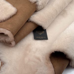 May include: Close-up of folded sheepskin rugs. The rugs have a soft, fluffy texture in cream and tan colours. The underside of the rugs is a smooth, tan suede. A black label with gold text reads "IMPERO LONDON".