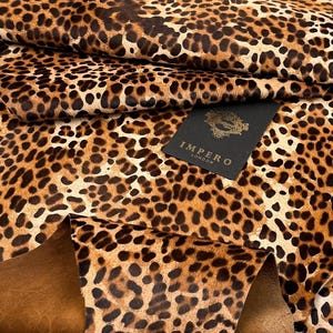Puede incluir: Primer plano de un trozo de cuero con estampado de leopardo. El cuero tiene un fondo beige con manchas marrones oscuras. Una etiqueta negra con texto dorado "IMPERO LONDON" es visible.