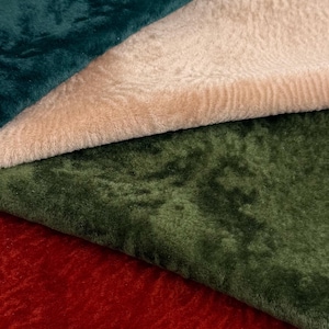 Op de afbeelding: Close-up van vier verschillende pluche stoffen in groen-, beige- en roodtinten. De texturen variëren van glad tot licht getextureerd, met een zachte, fluwelen uitstraling. De stoffen zijn gelaagd en tonen hun verschillende kleuren en texturen.